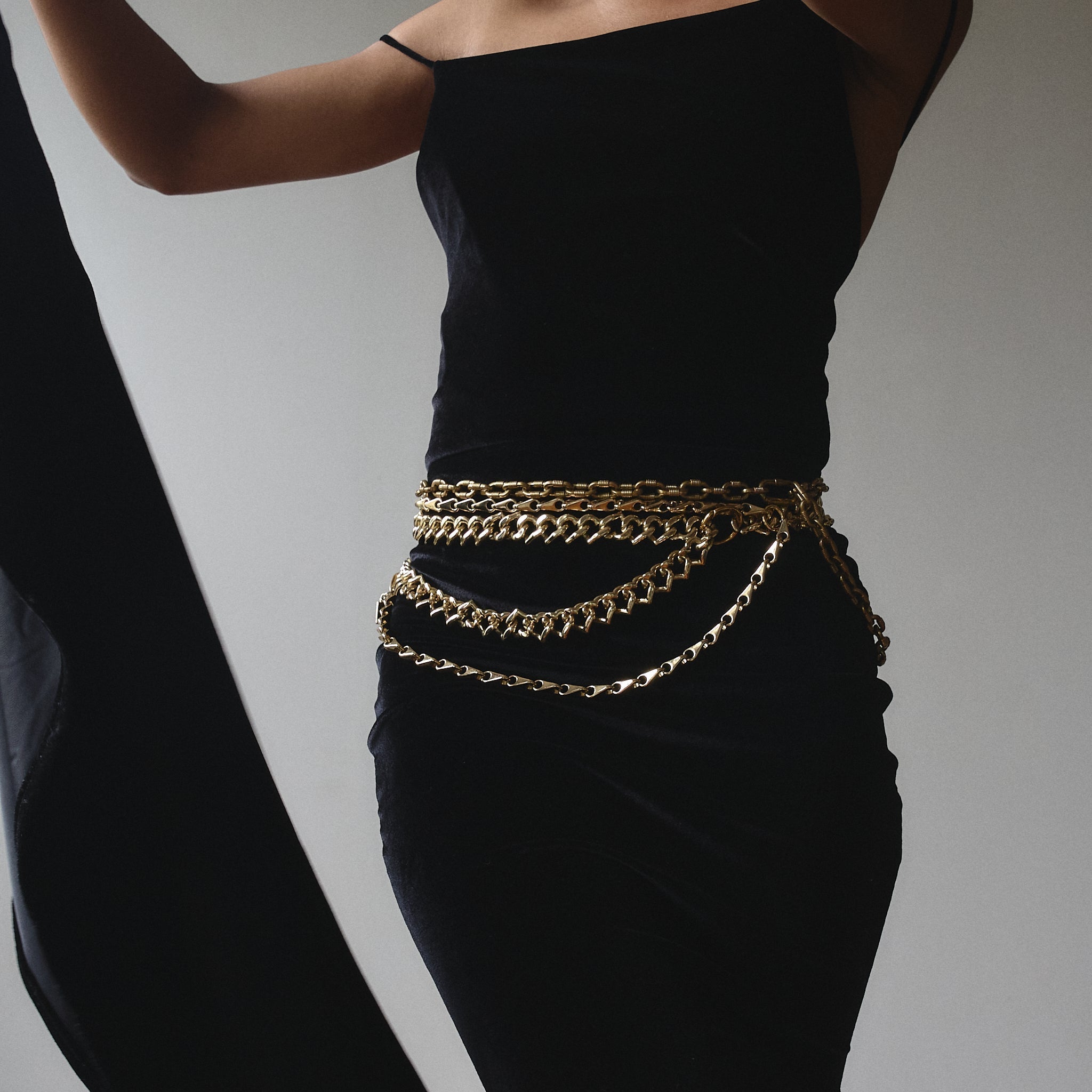 A woman wearing a black dress is cinched at the waist with a layered gold chain belt. The belt features multiple strands of varying chain styles, creating a draped effect around her waist.