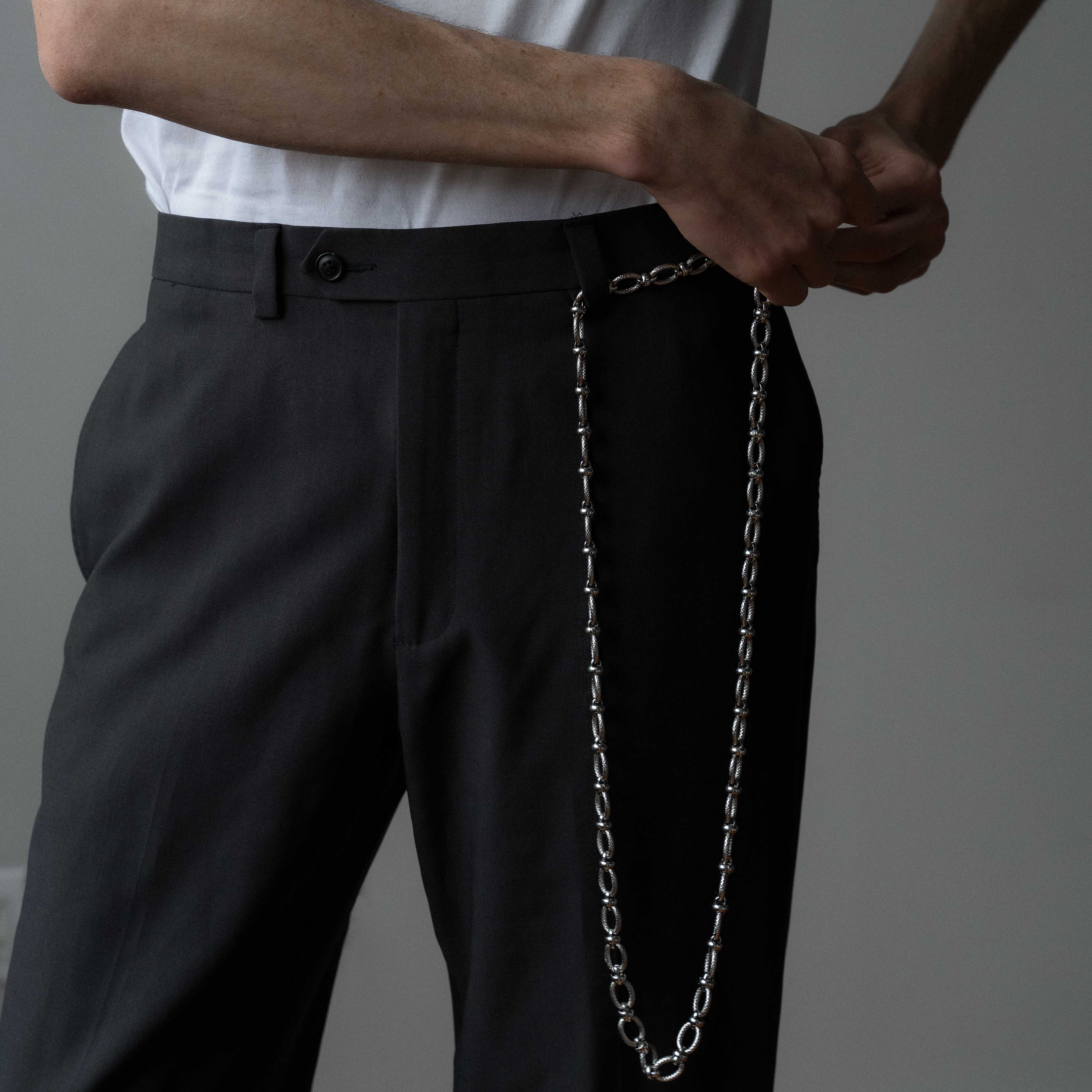 A photo of a man wearing a white t-shirt and black pants, stands in front of a mirror adjusting a silver chain belt at his waist. The belt has a modern, industrial design with a vintage touch.