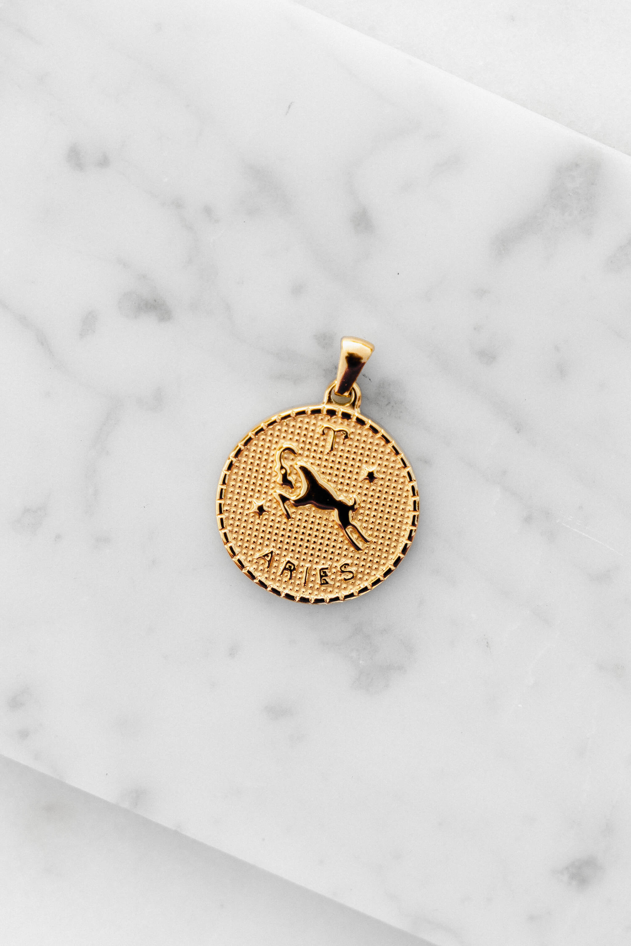 Aries zodiac sign gold coin charm laying on a white marble