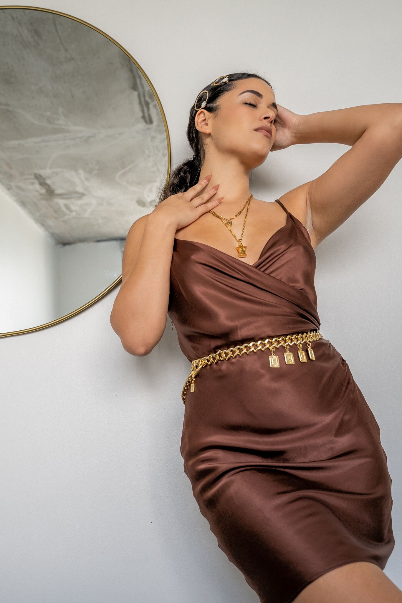Model in brown silky dress wearing Gold Lapo charm chain belt by the mirror on the wall