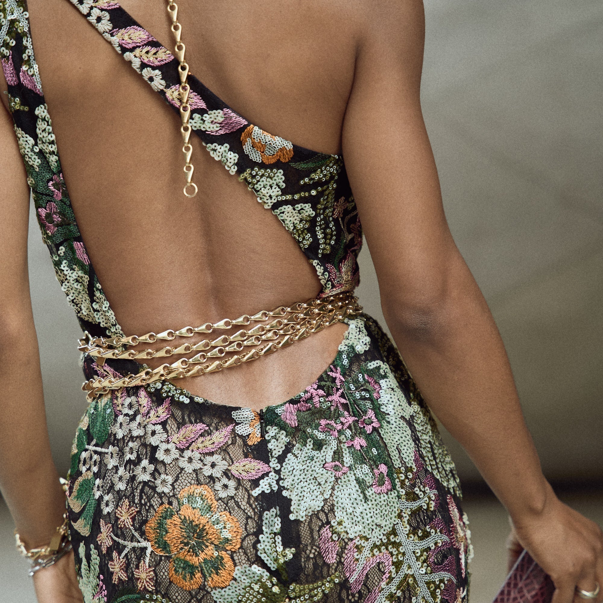 Close-up back view of a model in an open-back embellished floral gown styled with layered Lapo Lounge gold chain belts cinched at the waist, with a longer gold chain cascading from the neckline down the spine of the open back.