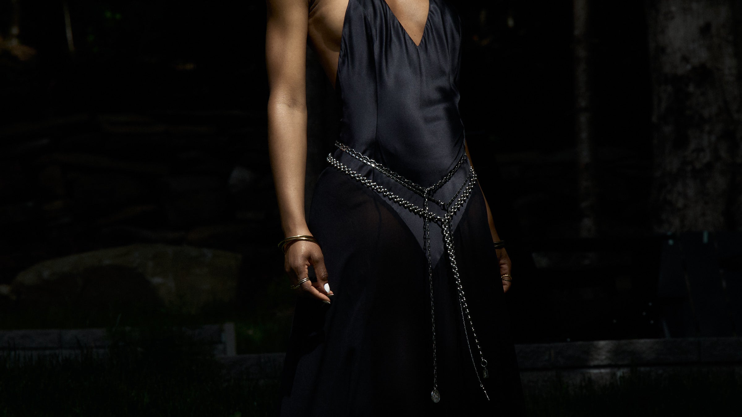 Woman wearing a black silk slip dress styled with layered silver chain belts against a dark background, showcasing minimalist, genderless jewelry from Lapo Lounge.